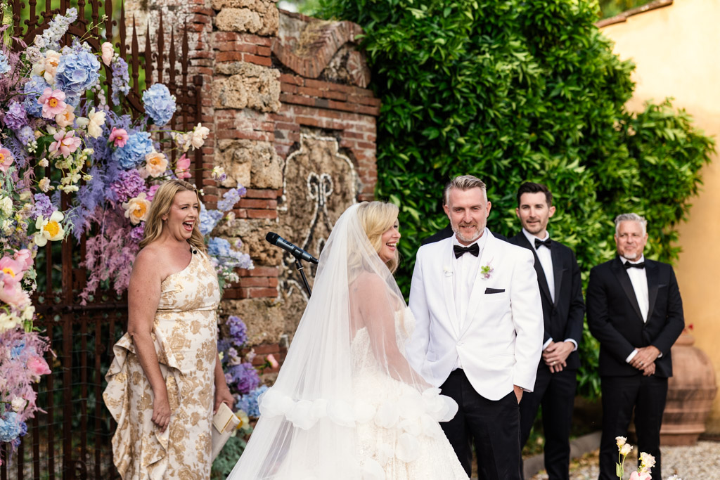 Bride and groom smile at each other during their outdoor Italian Villa wedding ceremony, surrounded by guests and vibrant flowers