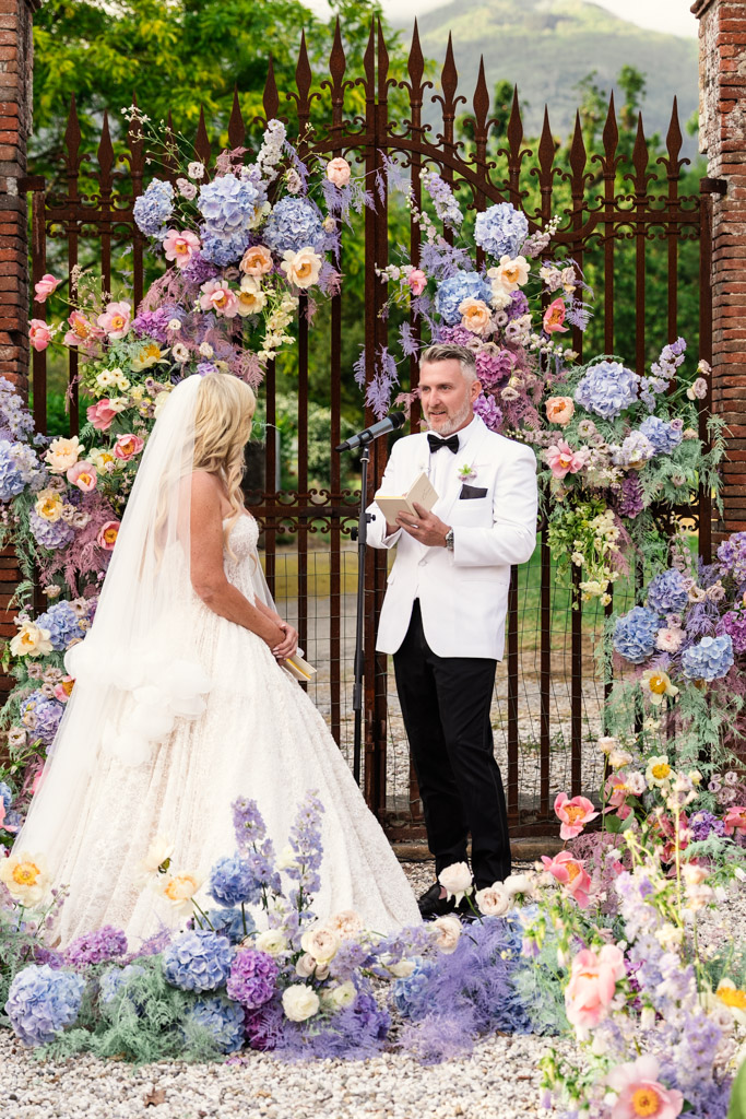 Bride and groom in front of a gate covered in flowers, exchanging vows during their Italian Villa wedding ceremony