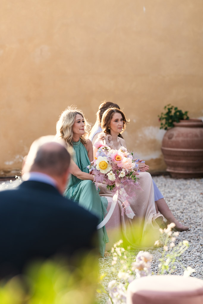 Two women sit outside during oan Italian Villa wedding; one holds a bouquet of pastel flowers
