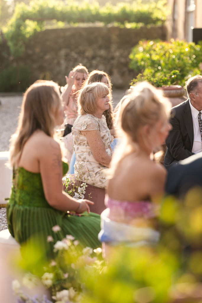 Guests sit outdoors in sunlight, surrounded by greenery, attentively watching an Italian Villa wedding ceremony