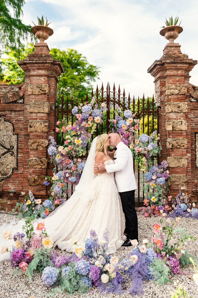 A bride and groom kiss in front of a floral arch and brick gate at their Italian Villa wedding celebration