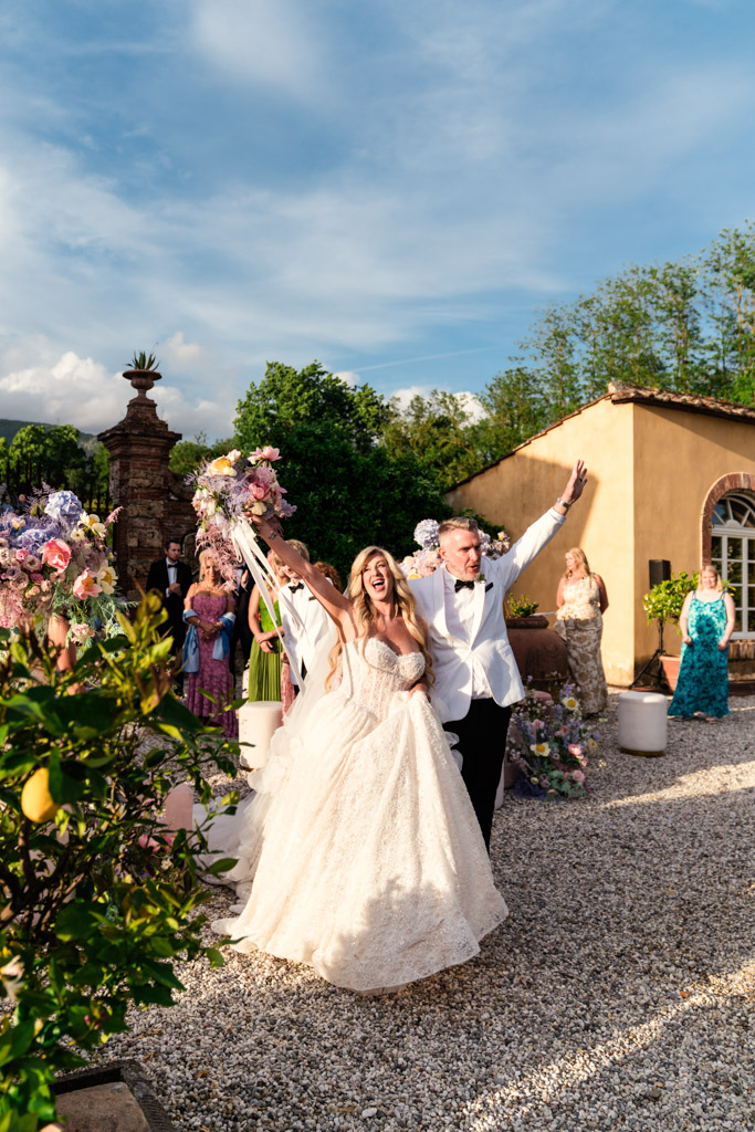 Bride and groom celebrate outdoors at their enchanting Italian Villa wedding, smiling and raising hands, surrounded by guests and flowers