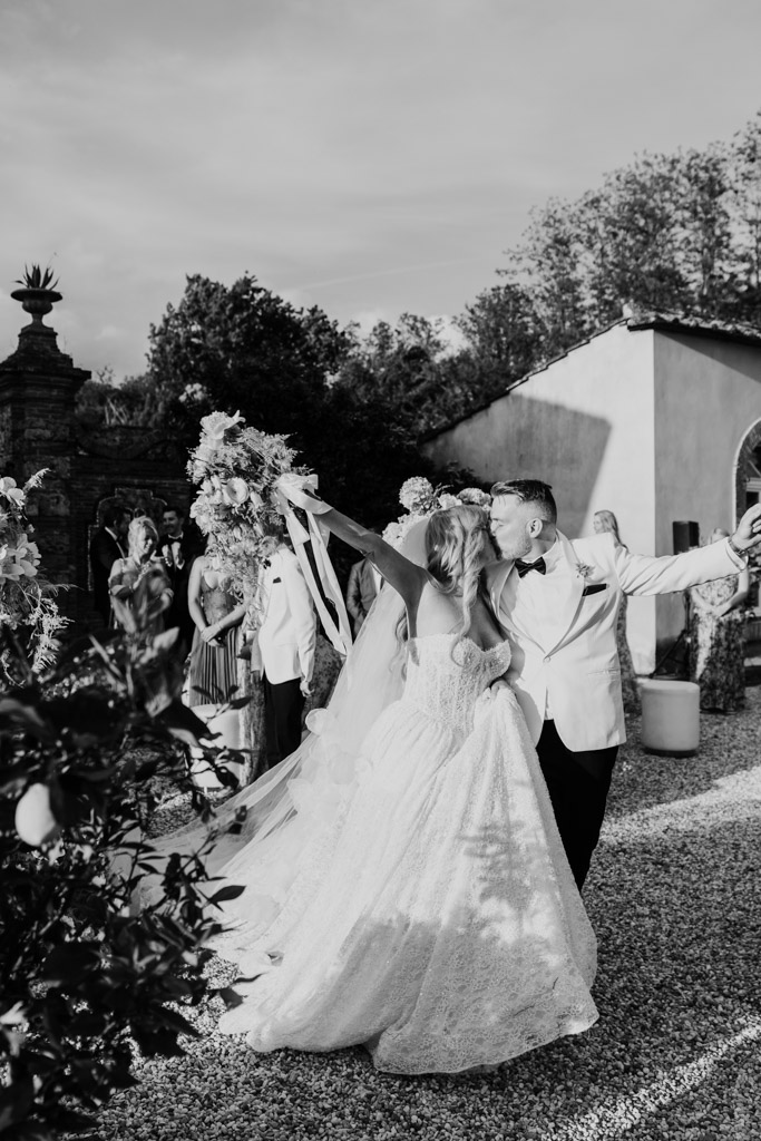Blacka dn white photo of bride and groom outdoors at their Italian Villa wedding, kissing and the bride holding a bouquet, with guests and lush greenery in the background