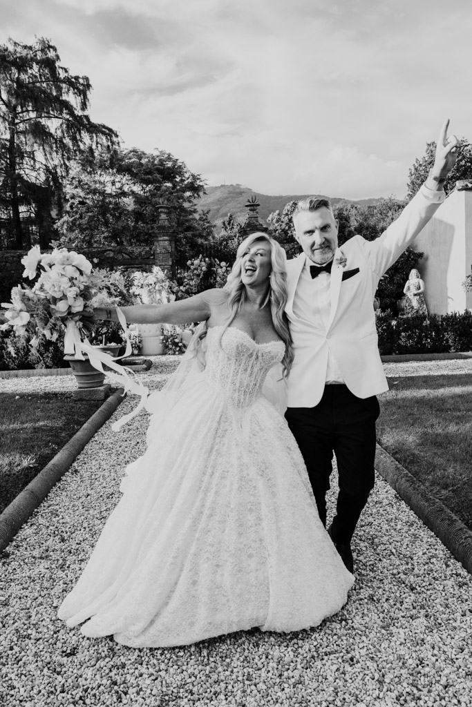 Black and white photo of bride and groom joyfully posing outdoors at their Italian Villa wedding, arms raised and smiling