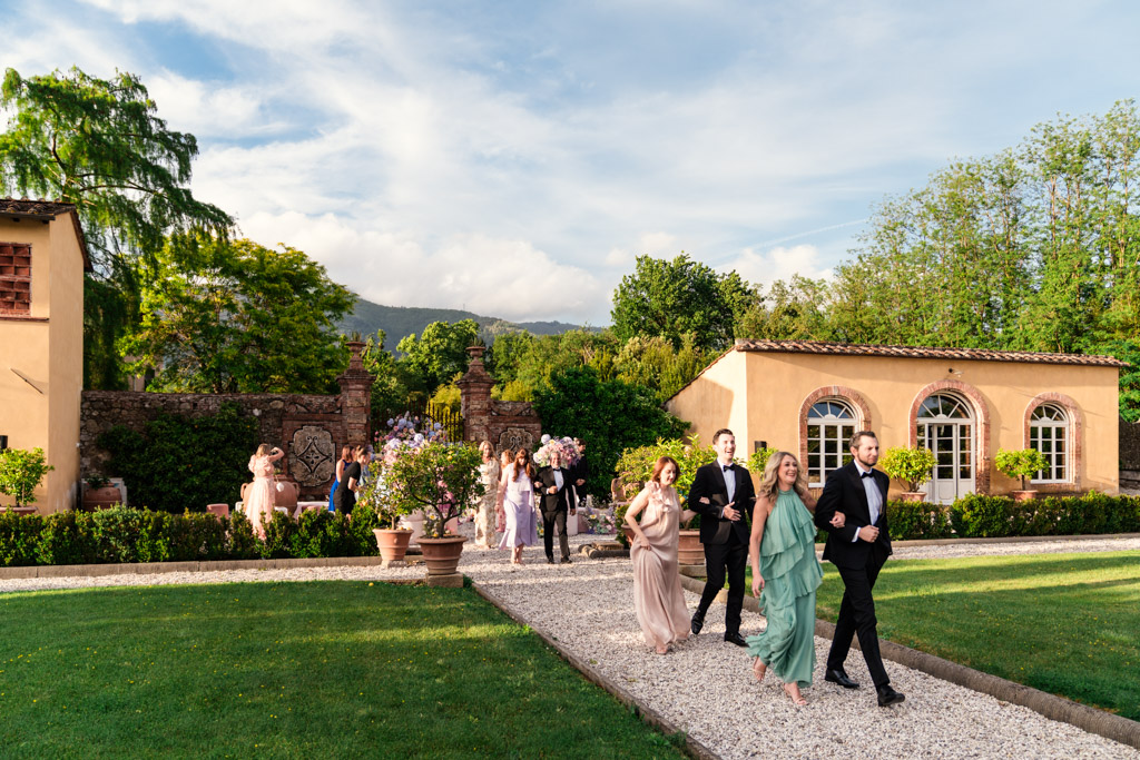 Wedding guests in formal wear walk along a garden path near rustic buildings and lush greenery during an Italian Villa wedding celebration