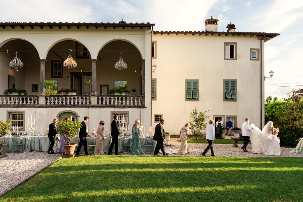 A wedding party walks in line outside a grand Italian Villa wedding venue in Tuscany, surrounded by elegant arches, glittering chandeliers, and set dining tables