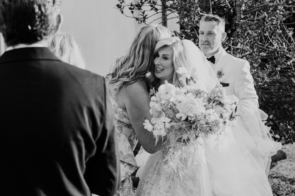 Black and white photo of bride in a white dress hugging a woman while holding a bouquet at an elegant Italian Villa wedding; the groom stands behind them, with guests gathered in the foreground