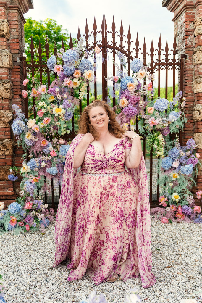 Smiling woman in a pink floral dress stands before a gate decorated with colorful flowers outdoors for an Italian Villa wedding celebration