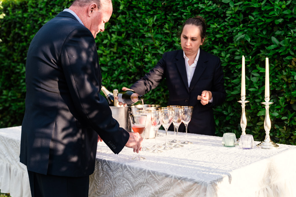 A server pours champagne into glasses at an outdoor table, candles flickering and lush greenery behind them, during Italian Villa wedding celebration