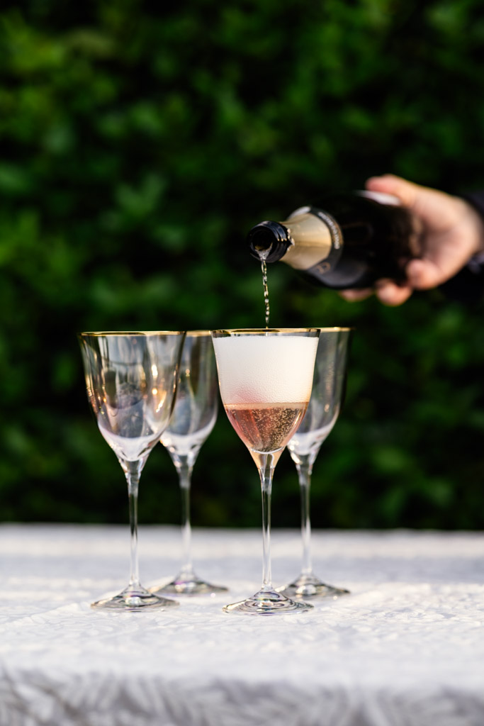 A hand pours sparkling wine into a glass beside two empty glasses on a white table outdoors, during Italian Villa wedding celebration