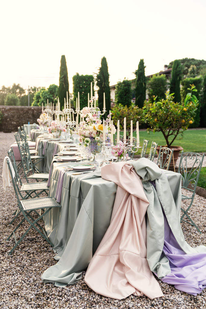 Outdoor table with pastel cloths, candles, flowers, and green chairs on a gravel path in a garden for Italian Villa wedding celebration