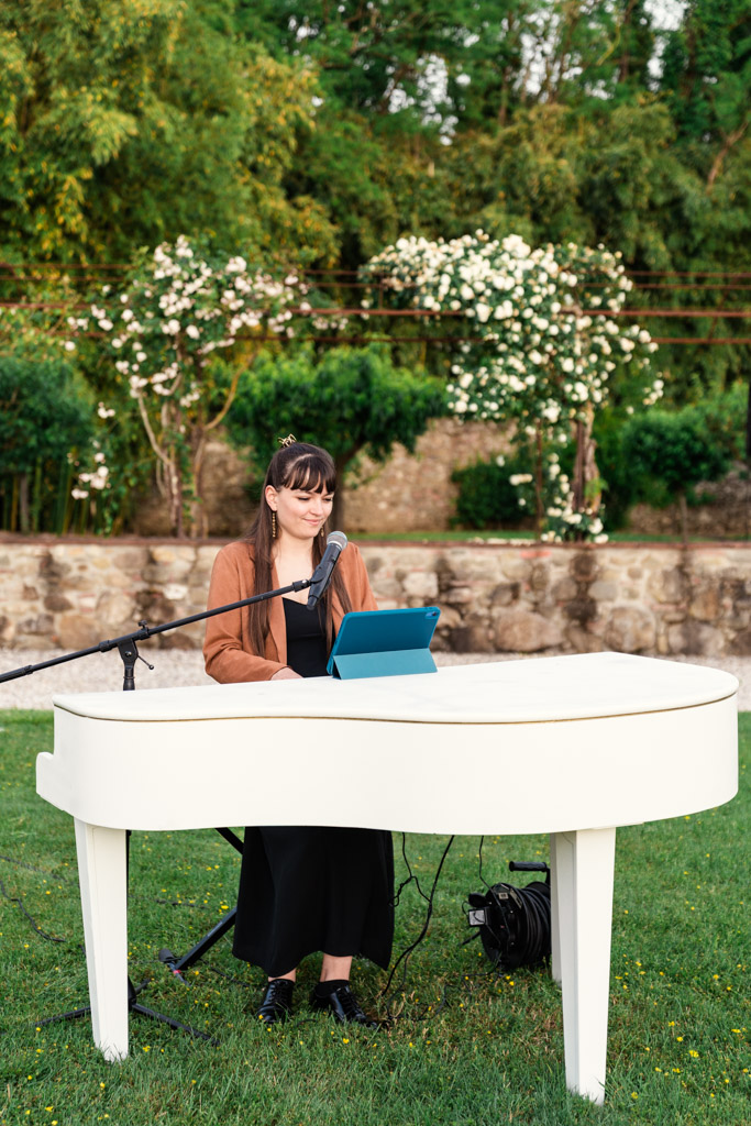 Woman playing a white piano outdoors at an Italian Villa wedding, with green trees with white flowers behind her