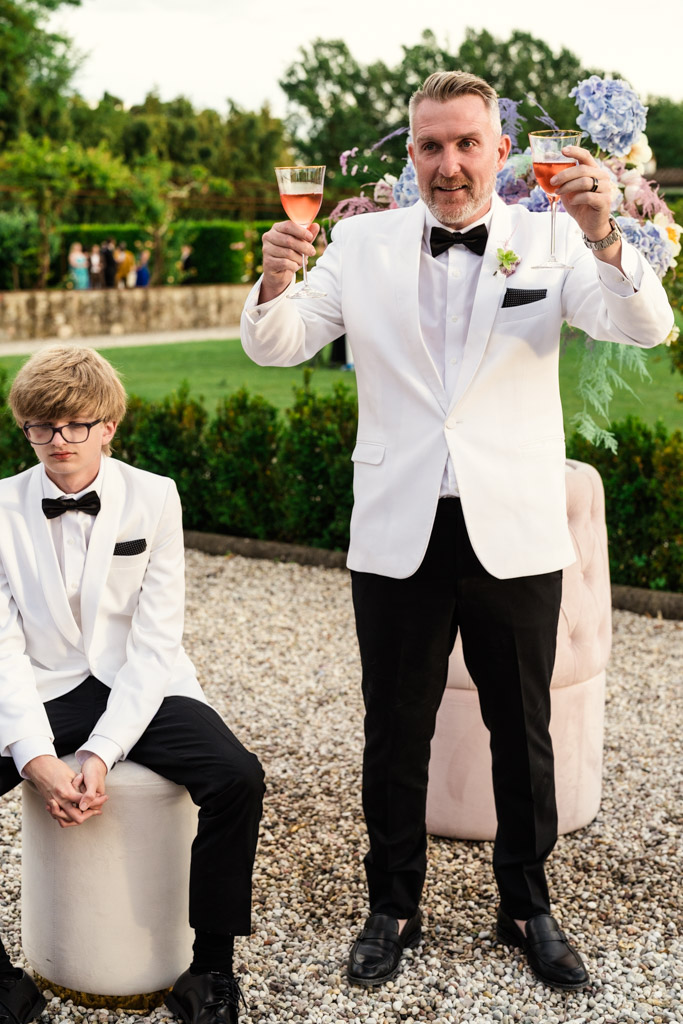 Two people in white tuxedos at an ; one sits with a neutral expression, Groom in white tuxedo holds up two drinks and smiles with a groomsman seated nearby during his Italian Villa wedding reception