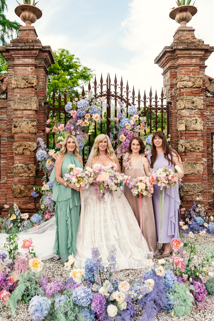Bride and bridesmaids in pastel dresses pose with bouquets before a flower-adorned gate and brick pillars during Italian Villa wedding celebration