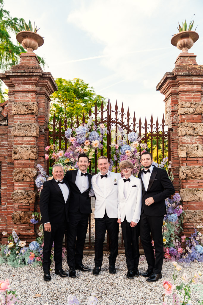 Groom and groomsmen in tuxedos stand in front of a floral-decorated gate at an outdoor Italian Villa wedding