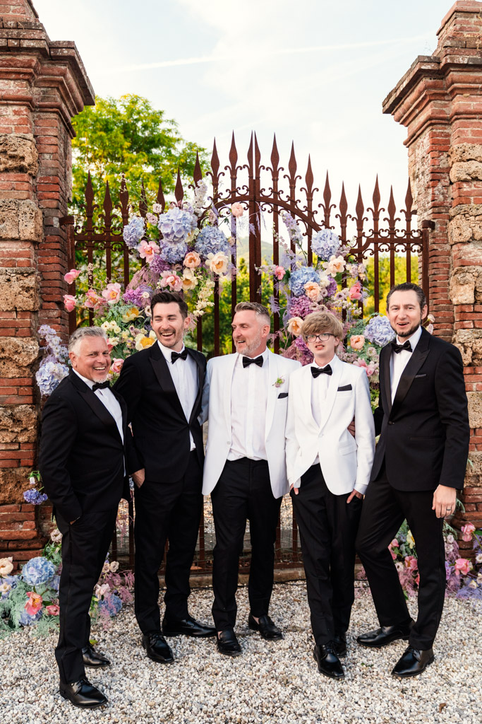 Groom and groomsmen in tuxedos stand and smile in front of a floral-decorated iron gate and stone archway during Italian Villa wedding celebration