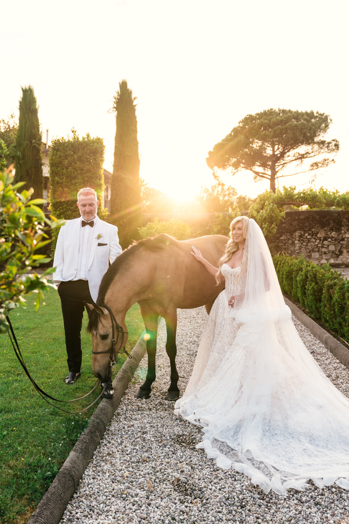 Bride and groom stand with a horse on a garden path at sunset, the bride wears a long veil, during their Italian Villa wedding celebration