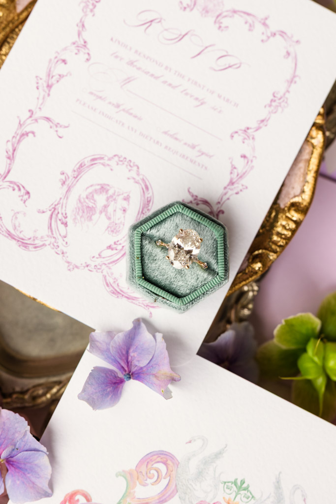 An engagement ring in a green box rests on RSVP cards, surrounded by delicate flowers and a gold tray in the background for Italian Villa wedding