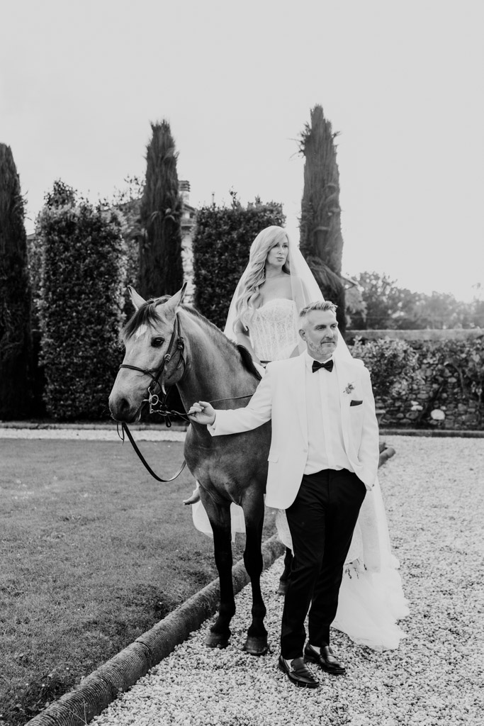 Black and white photo of bride in a wedding dress on a horse, led by groom in a white suit, outside in a Italian Villa garden