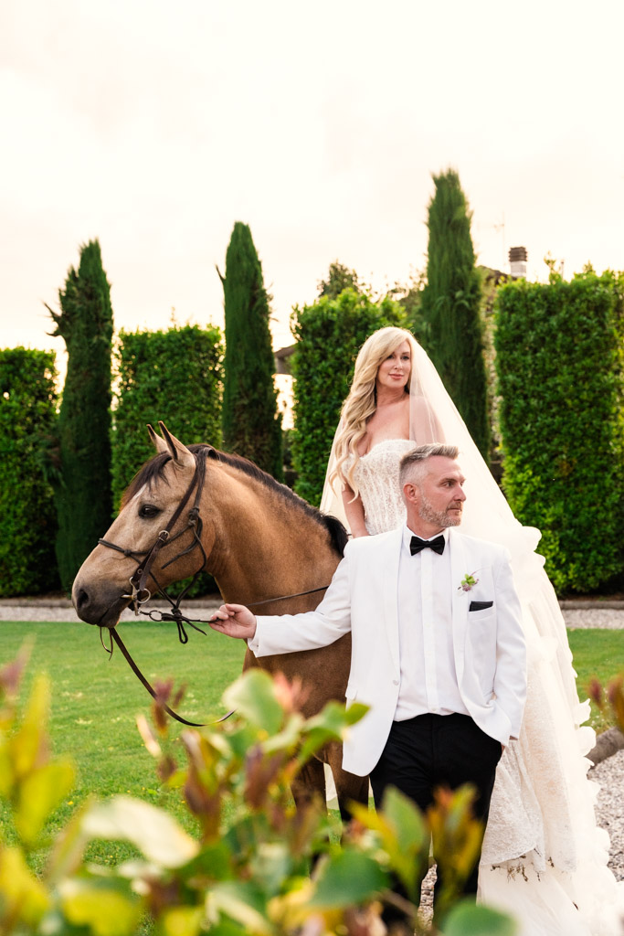 Bride on a horse in wedding dress, groom in white tuxedo leads the horse through a lush garden at an Italian Villa