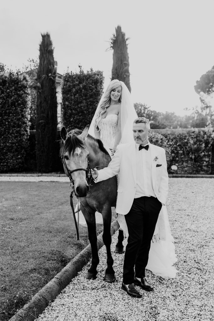 Black and white photo of bride in a wedding dress on a horse, led by a groom in a suit on a gravel path outdoors during their Italian Villa wedding celebration