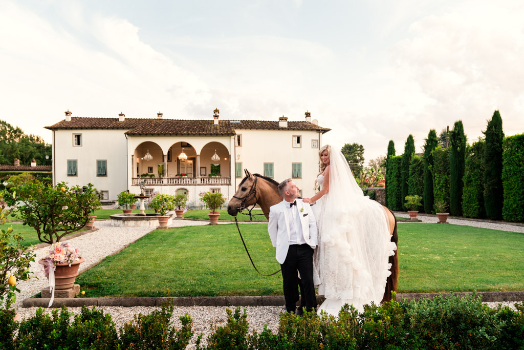Bride on a horse smiles at groom in white suit, with an elegant Italian Villa wedding and lush garden in the background