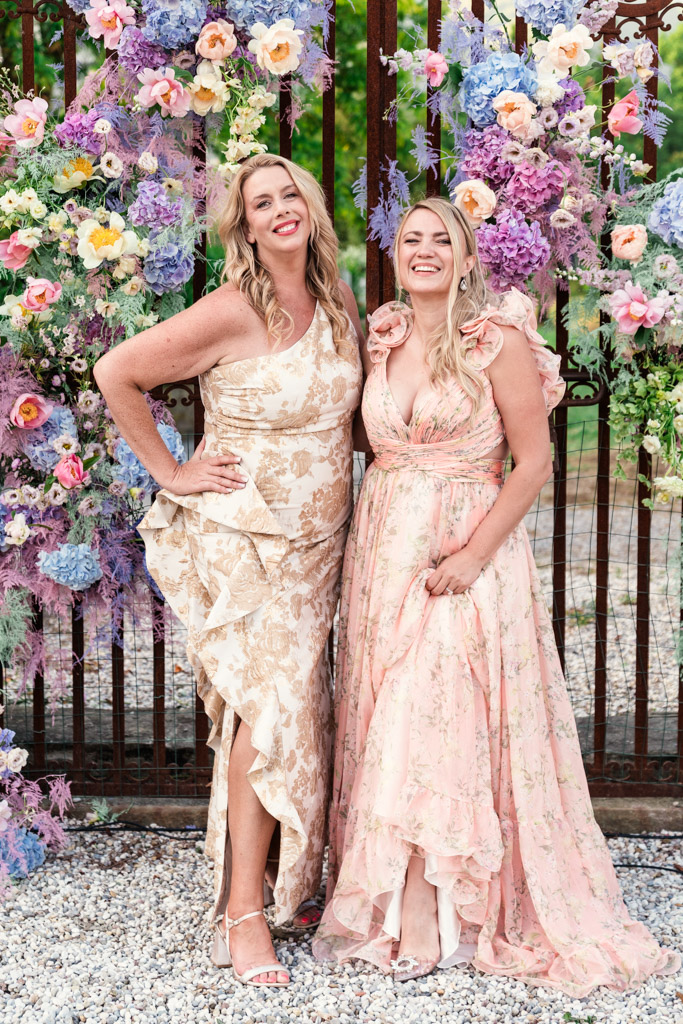 Two women in floral dresses smile together in front of a colorful floral backdrop and a metal gate during Italian Villa wedding celebration