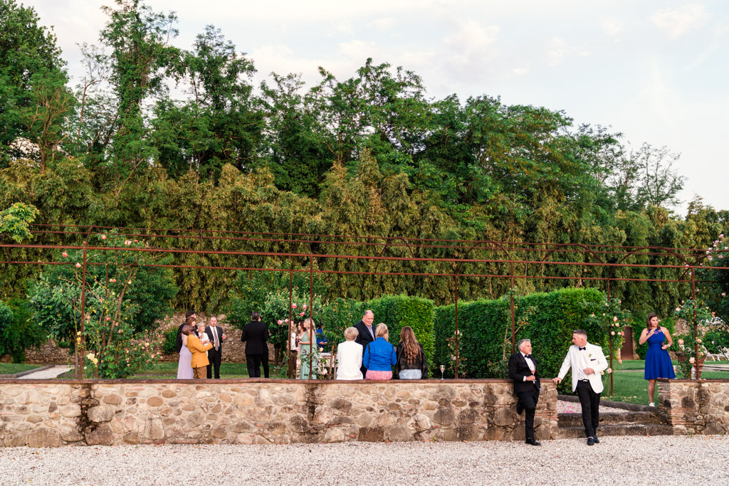People dressed formally gather outdoors by a stone wall and lush greenery, under a partly cloudy sky during an Italian Villa wedding celebration