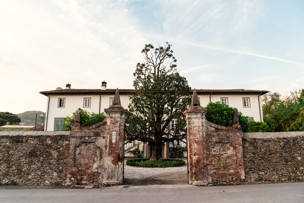 Italian Villa brick gate with stone pillars opens to a large white house and a tall tree in the center courtyard