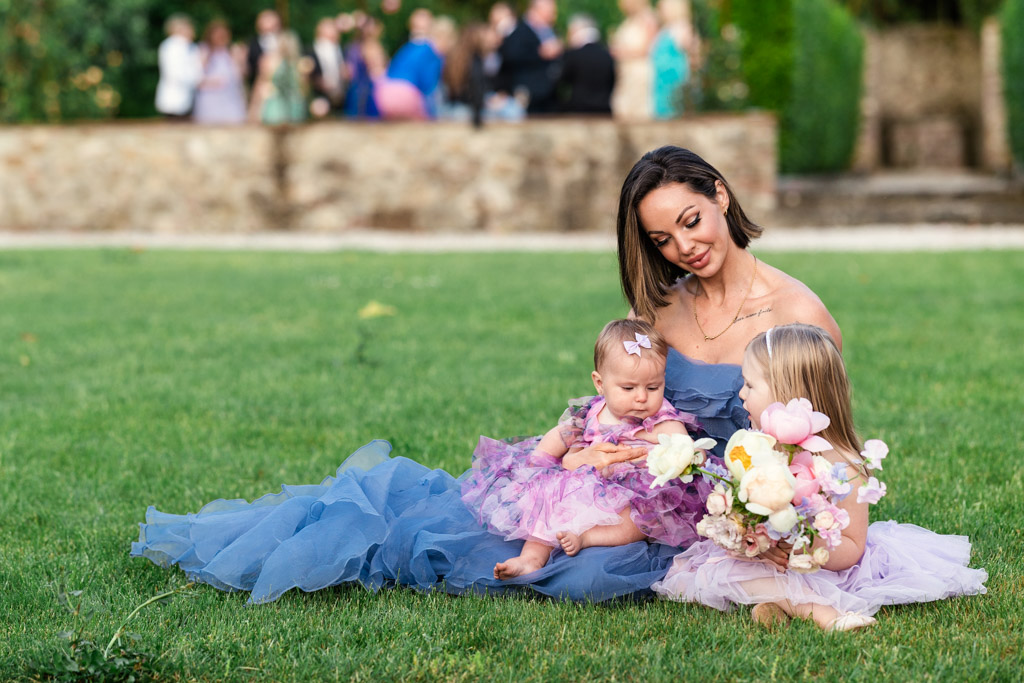 Woman in a blue dress sits on the grass with two young girls in purple dresses holding flowers, capturing a charming moment at an Italian Villa wedding