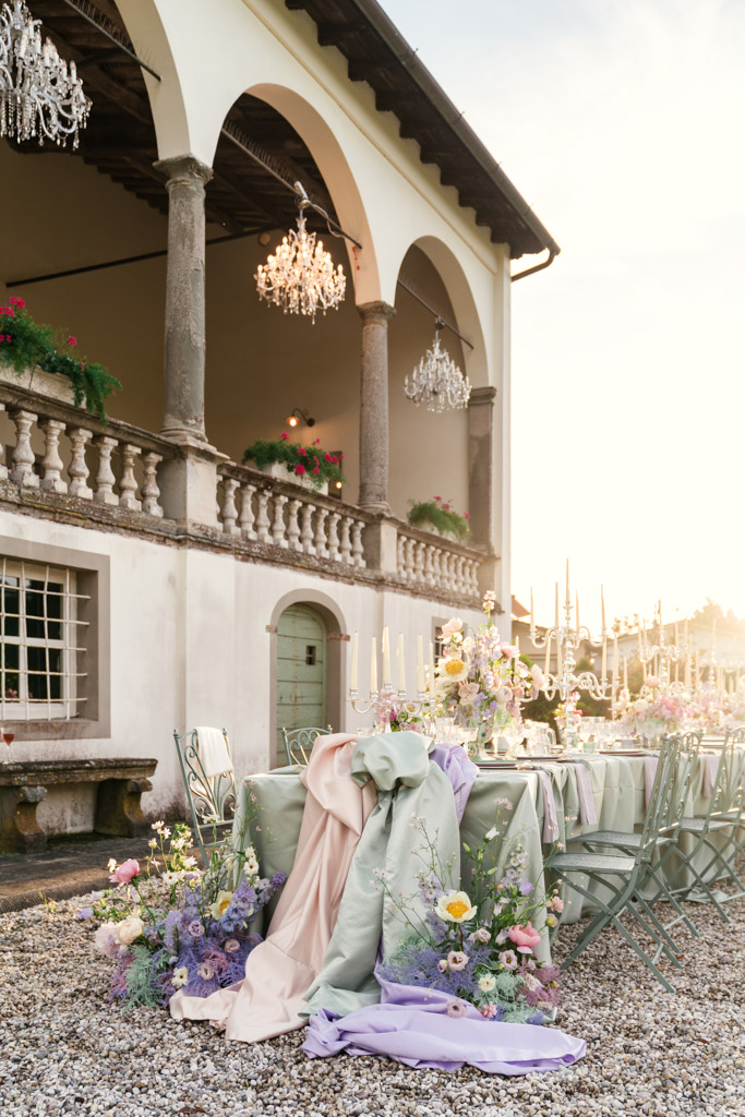 Outdoor table setup with pastel linens and flowers beside a grand building with chandeliers and columns for an Italian Villa wedding