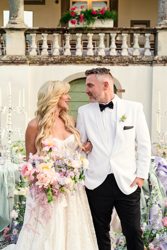 Bride and groom stand arm in arm outdoors at their Italian Villa wedding, smiling at each other, holding a bouquet of pastel flowers