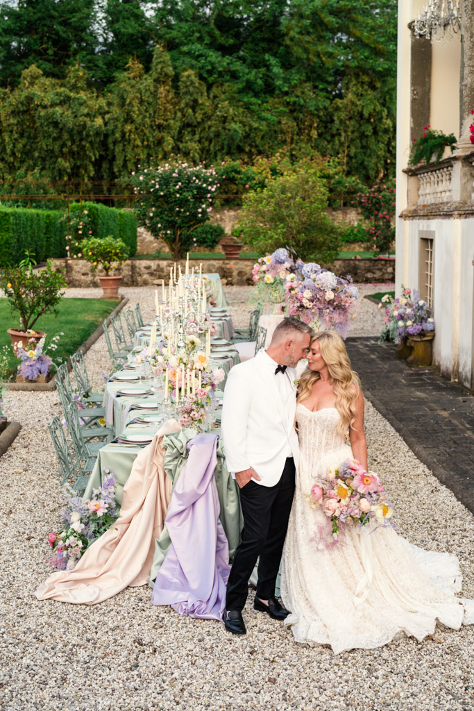 A bride and groom stand by a pastel wedding table outdoors at their Italian Villa wedding, surrounded by flowers and candles