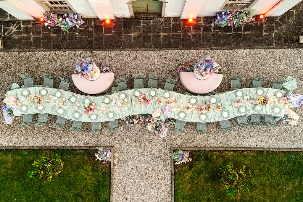 Overhead view of outdoor banquet table set for many guests with pastel decor, flowers, and green chairs for an Italian Villa wedding celebration