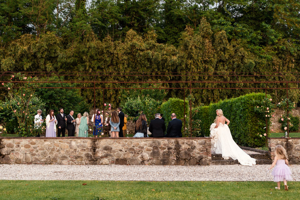 A bride walks across a stone terrace at her Italian Villa wedding towards a group of people as a young girl stands in the foreground