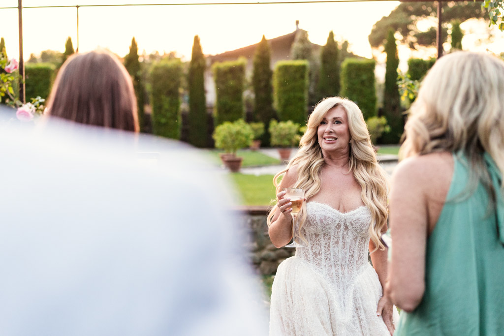 Bride in a white dress holding a drink, smiling outdoors at an Italian Villa wedding, with guests and lush greenery in the background