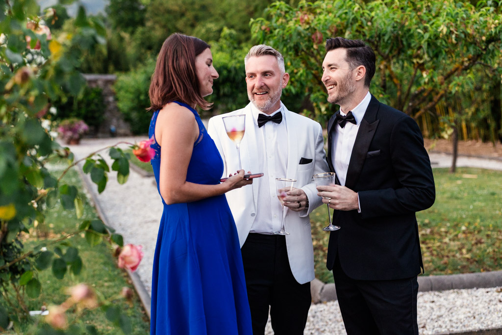 Groom and two guests in formalwear stand outside, smiling and talking, holding drinks in an Italian Villa garden