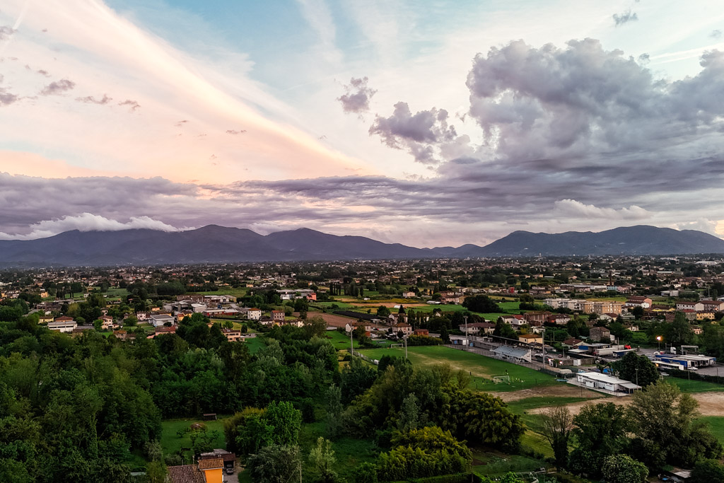 Town in Tuscany with green trees, buildings, and distant mountains under a colorful, partly cloudy sky at sunset