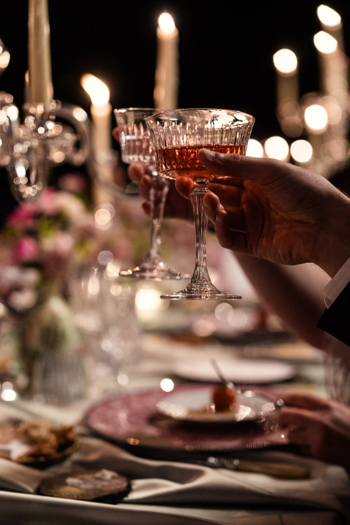 Hands raising champagne glasses in a toast at a candlelit dinner with elegant table settings during Italian Villa wedding reception