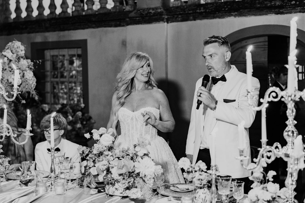 Black and white photo of bride and groom stand and speak at their elegant, flower-filled Italian Villa wedding reception