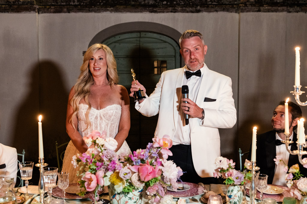 Groom in a white tux holds a trophy and microphone beside bride in a white dress at a candlelit Italian Villa wedding reception