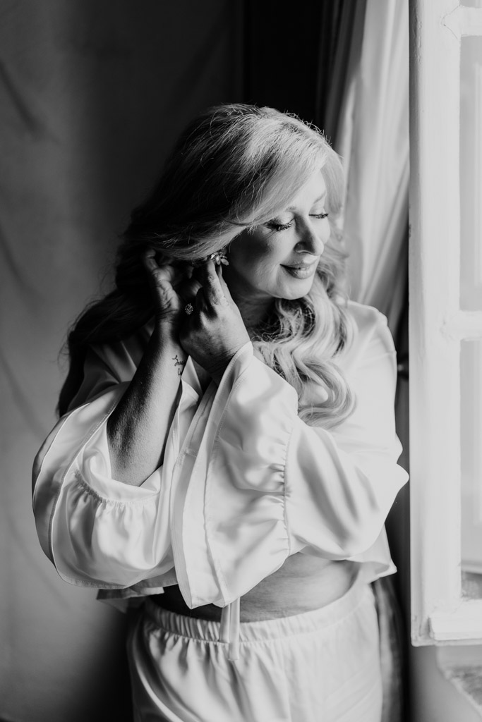Black and white photo of bride in a silk robe putting on earrings, standing by a window with soft natural light, preparing for her Italian Villa wedding