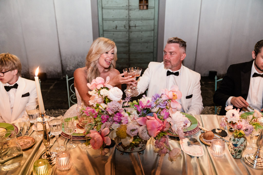 Bride and groom in white toast at a flower-filled wedding table, smiling, with guests in black tuxedos at Italian Villa wedding reception