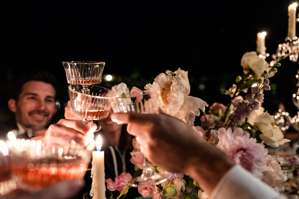 People clinking glasses at a candlelit Italian Villa wedding celebration, with flowers and candles on the table, smiling faces visible