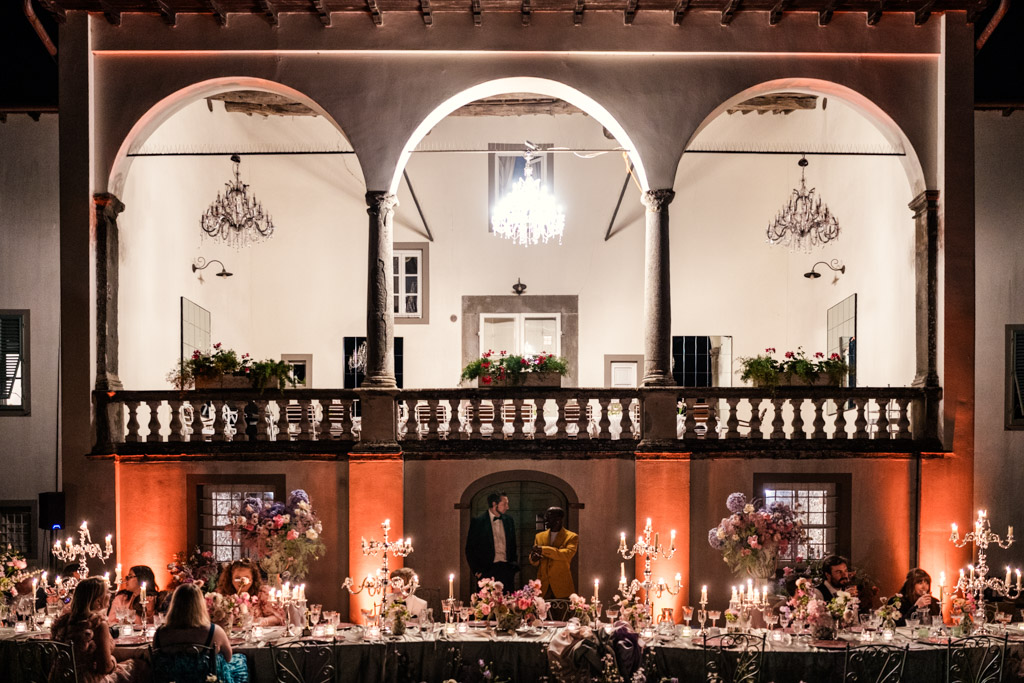 Elegant candlelit dinner outdoors with guests seated under a balcony at night, surrounded by chandeliers and flowers during Italian Villa wedding celebration