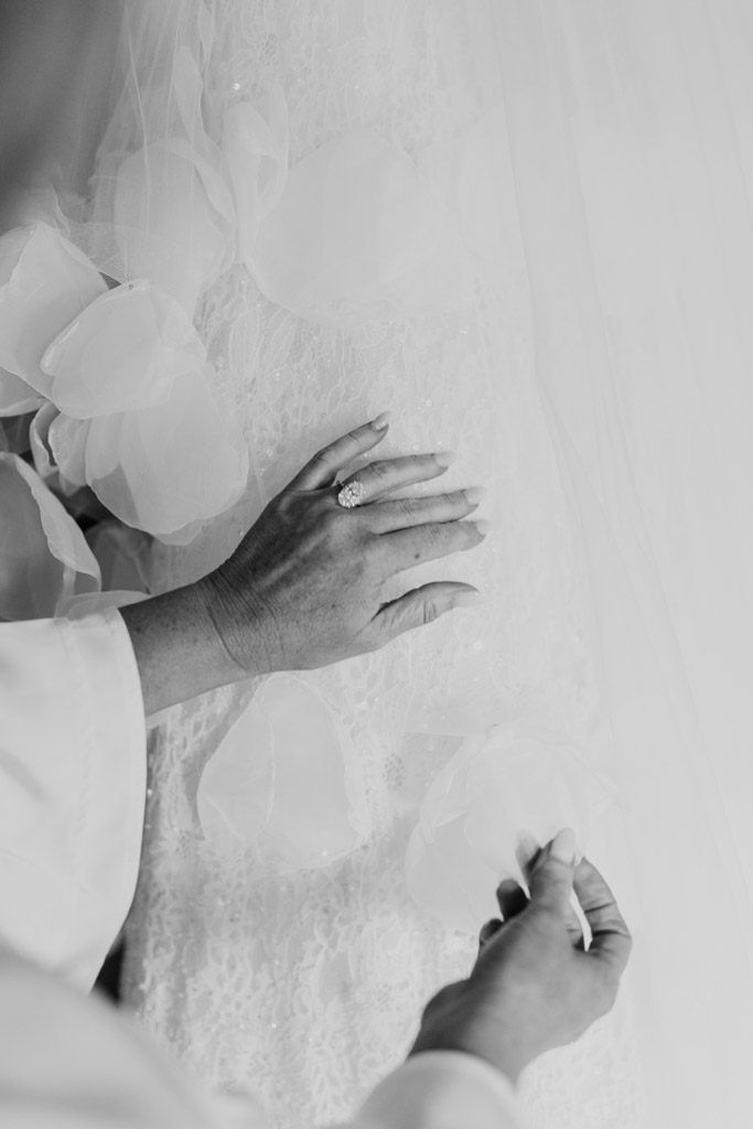Hands adjusting delicate floral lace, with a ring visible, on a white wedding dress in soft lighting