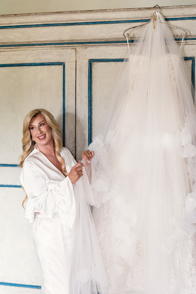 happy bride in a white robe holds a wedding dress hanging on a door with blue trim while getting ready for her Italian Villa wedding