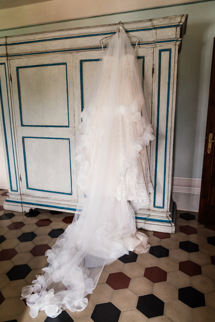 A white wedding dress with a long veil hangs on an antique wardrobe in a room with tiled floor, inside an Italian Villa
