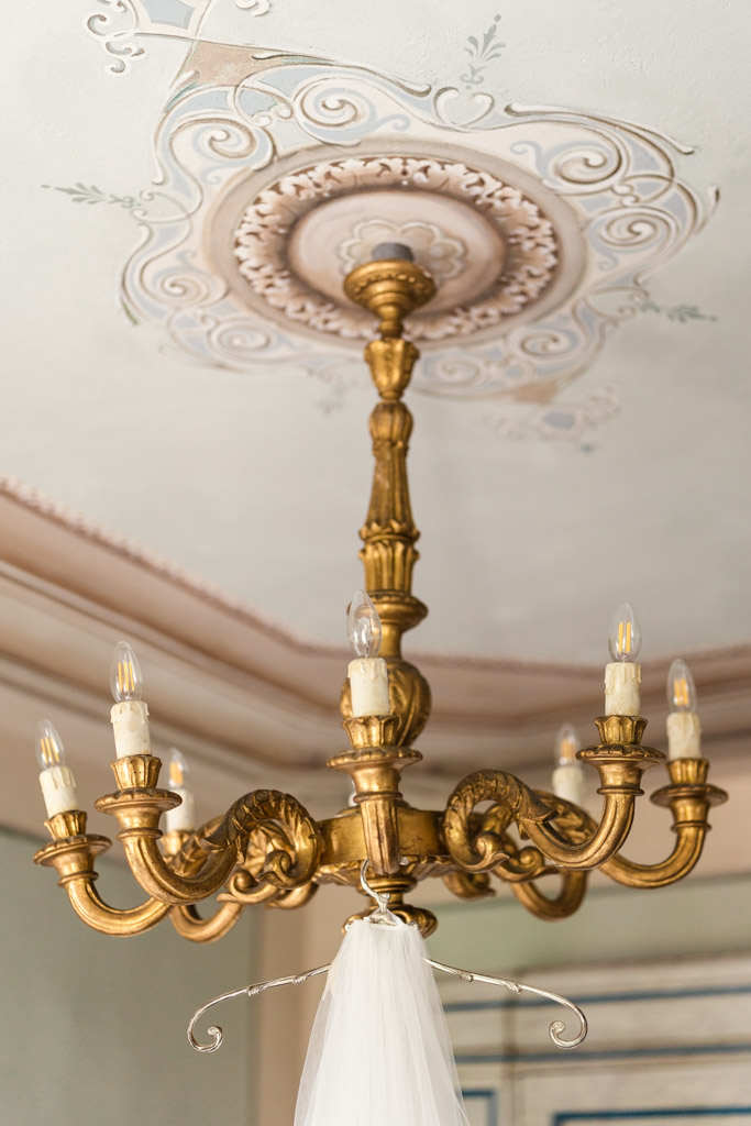 Gold chandelier with eight candle-like lights hanging from an ornate white ceiling with dress hanging from it