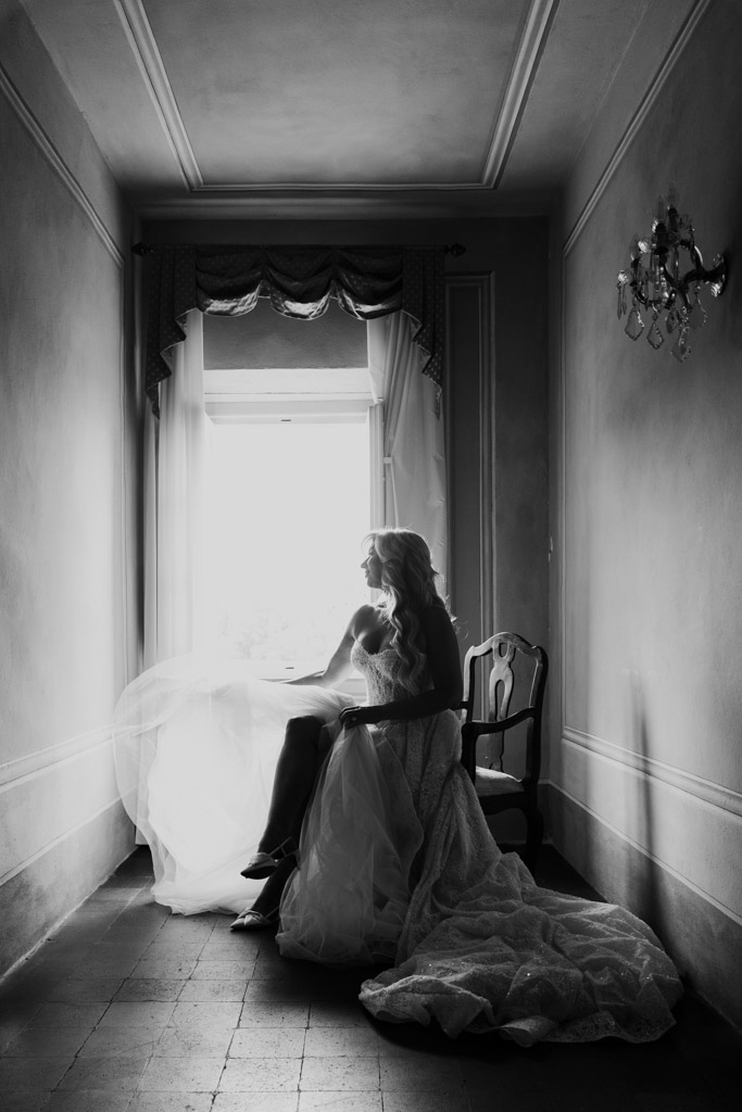 Black and white photo of bride in a gown by a bright window in a dark hallway, looking outside, while getting ready for her Italian Villa wedding